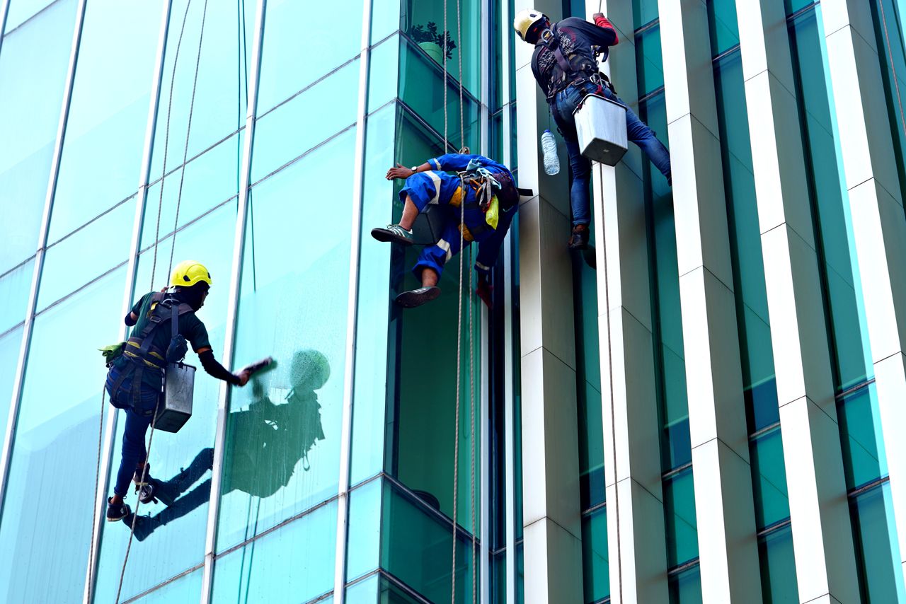 services-04 Three window cleaners suspended on ropes clean a modern glass building facade during the day.