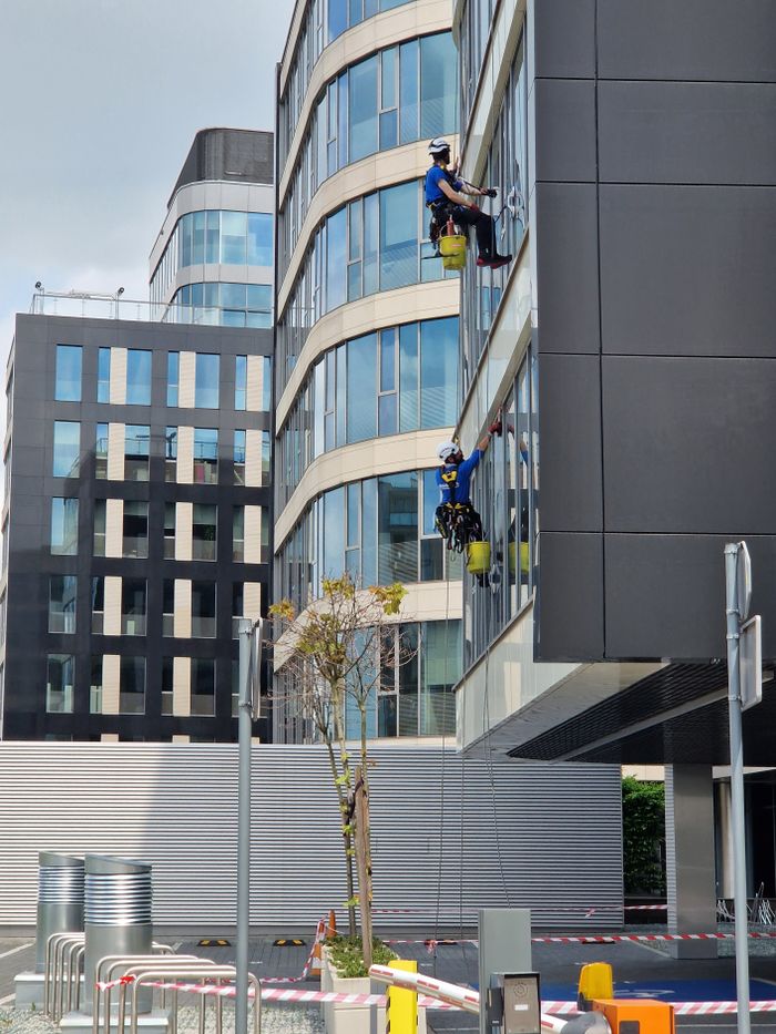 who-we-are Two workers clean windows on a tall office building in a city setting.